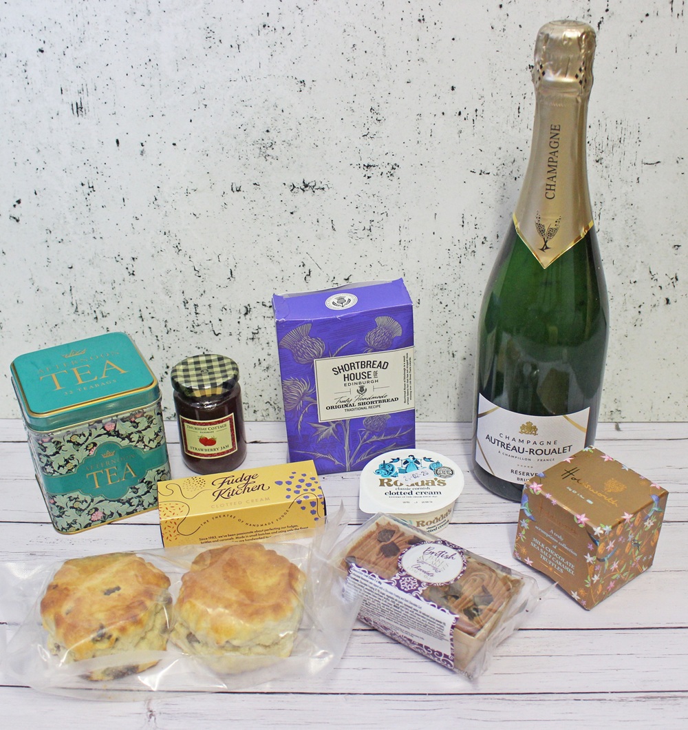 A selection of gourmet treats including a bottle of champagne, tea caddy, jar of jam, shortbread, fudge, scones, cheese, oatcakes, and a small floral box, all arranged on a light wooden surface.