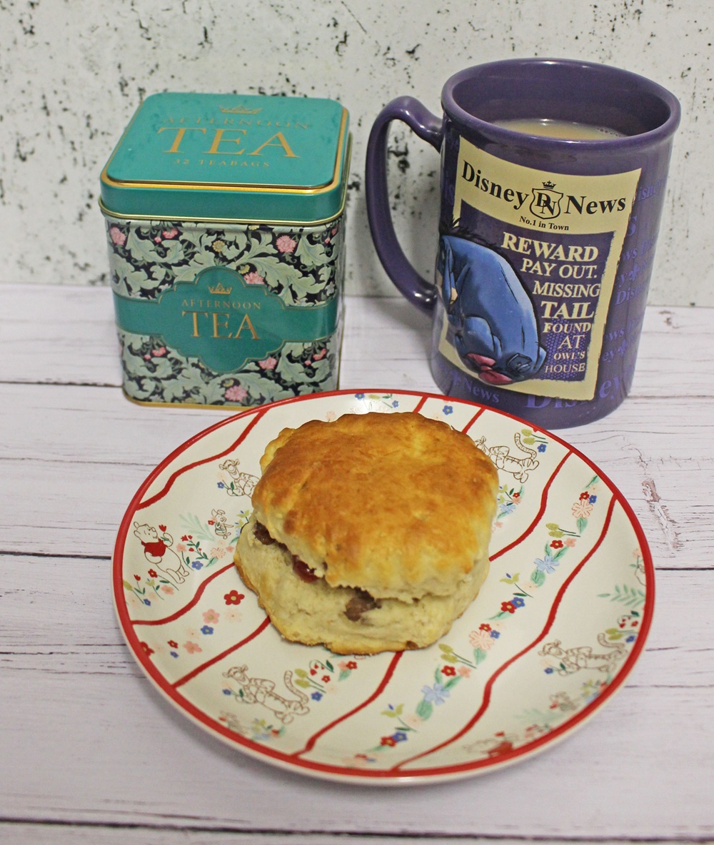 A plate with a scone, a blue floral tin labelled Afternoon Tea, and a mug with Disney's Eeyore and a Missing Tail reward poster, all on a white wooden surface.