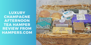 A wicker hamper filled with assorted treats, tea, and sweets sits on straw filler. Text on a blue background reads “Luxury Champagne Afternoon Tea Hamper Review from Hampers.com”.
