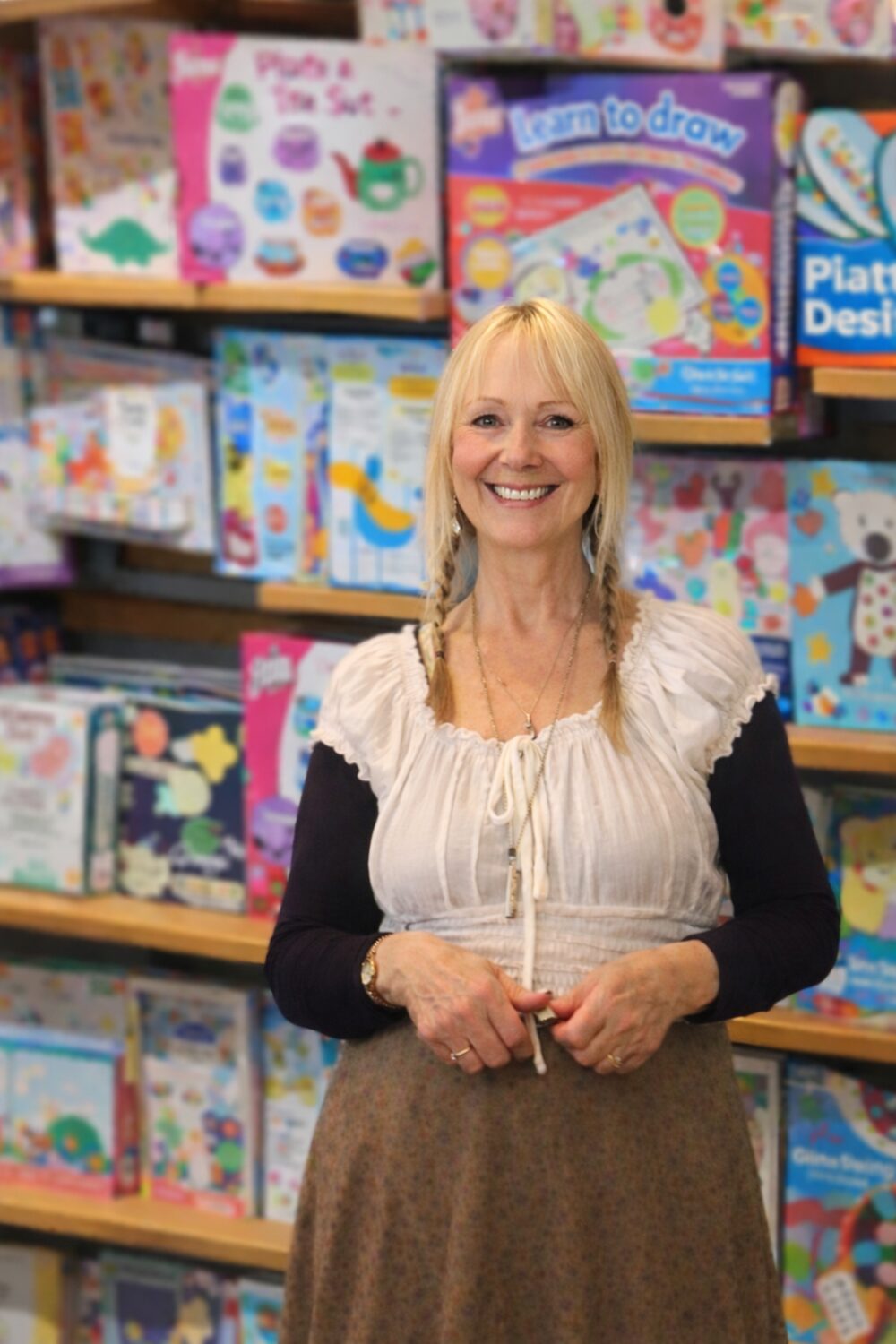 A smiling woman with long blonde hair stands in front of shelves filled with colourful children's art and craft kits. She is wearing a white blouse over a black shirt and a brown skirt.