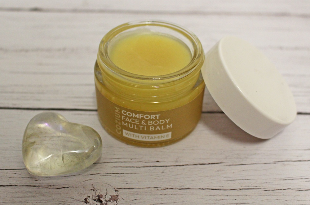 An open jar of yellow face and body multi-balm with vitamin E sits on a white wooden surface next to its white lid. A heart-shaped clear stone lies beside the jar.