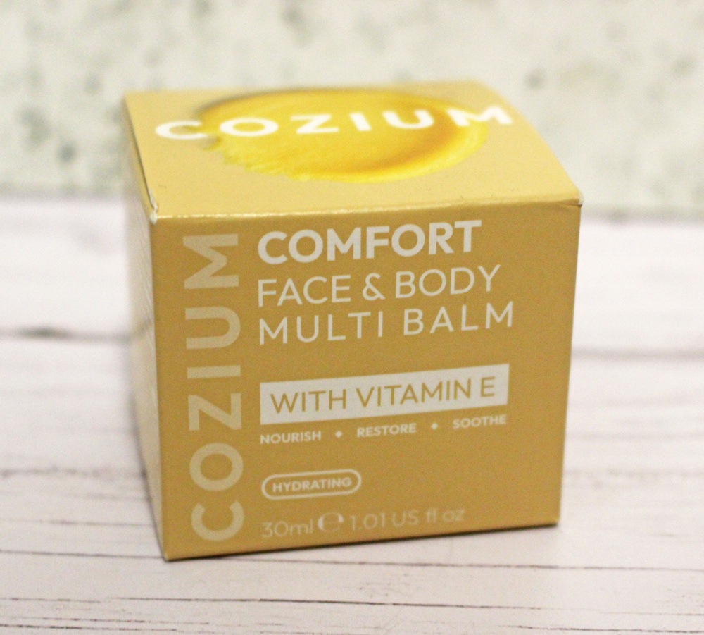 A yellow box of Cozium Comfort Face & Body Multi Balm with Vitamin E, hydrating formula, 30ml (1.01 fl oz). The box highlights benefits: nourish, restore, and soothe.