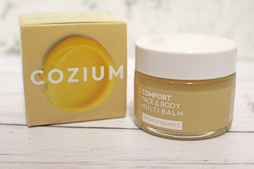 A jar and box of Cozium Comfort Face & Body Multi Balm with Vitamin E, displayed on a white wooden surface. The packaging is beige with white text and a yellow swatch graphic on the box.