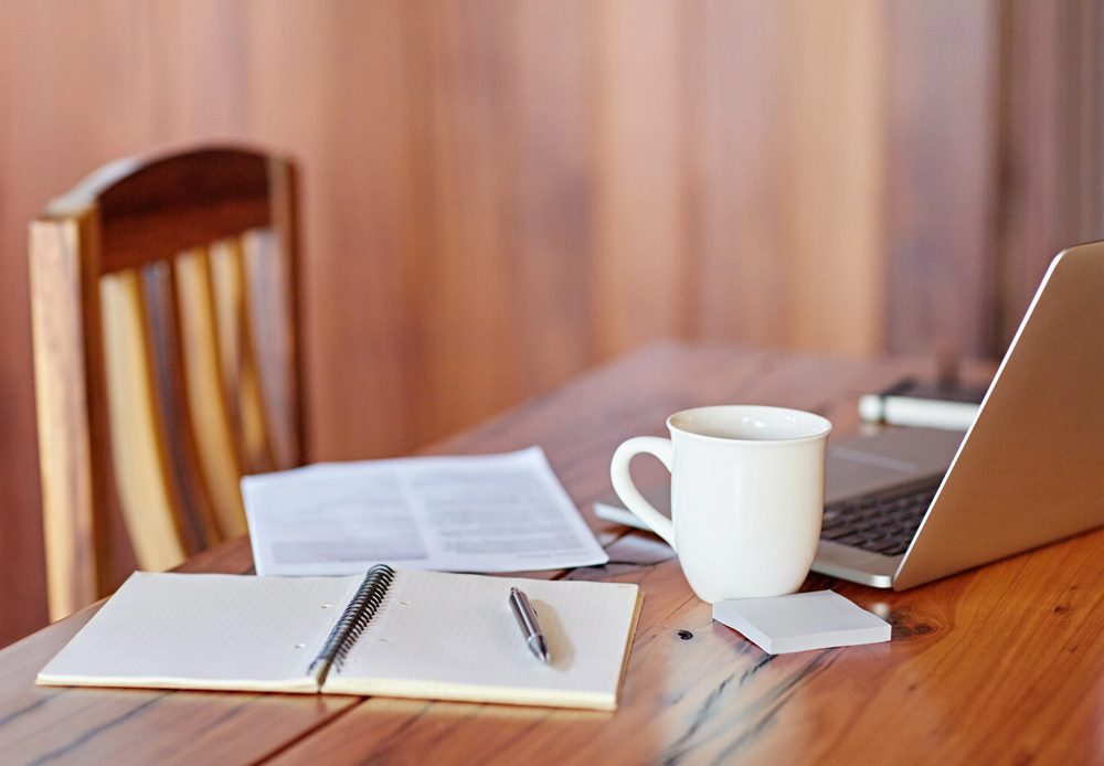 A wooden table with an open laptop, a white coffee mug, a spiral notebook with a pen, a stack of sticky notes, and a sheet of paper, with a wooden chair in the background.