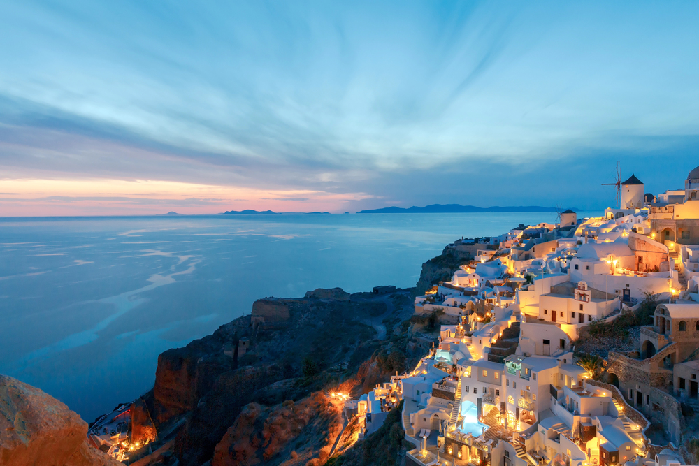 Whitewashed buildings with glowing lights on a hillside overlook a calm blue sea at sunset in Santorini, Greece. The sky is streaked with clouds, and distant islands are visible on the horizon.