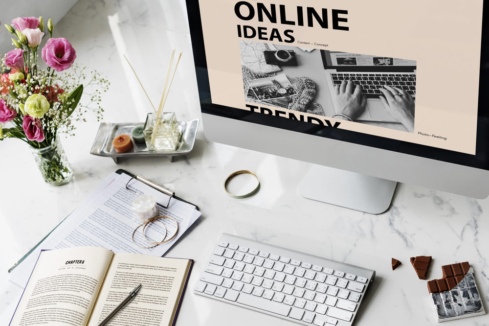 A desktop computer on a marble table displays a screen reading Online Ideas Trendy. The desk has an open book, a notebook, pen, glasses, flower vase, and pieces of chocolate.