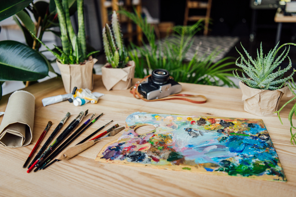 A wooden table with paintbrushes, a colourful artist’s palette, tubes of paint, an old camera, and several potted plants, creating a creative and natural workspace ambience.