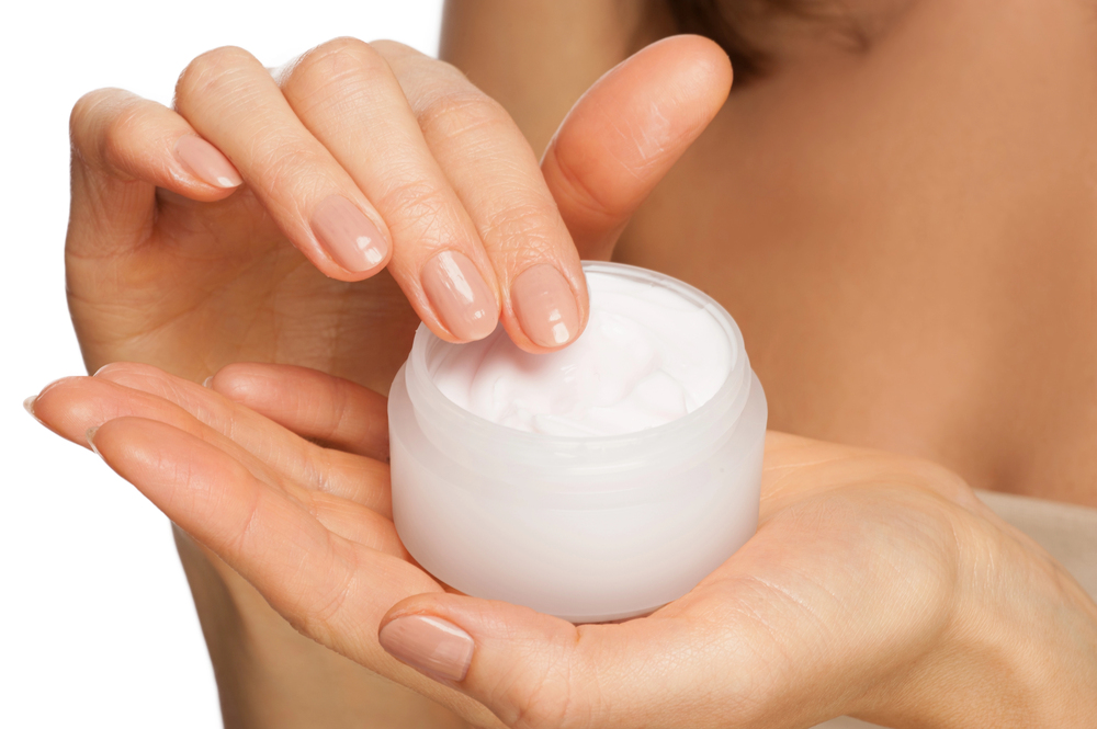 A close-up of a person’s hands holding an open jar of white cream. One hand is dipping a finger into the creamy substance, suggesting use of moisturising skincare or lotion.
