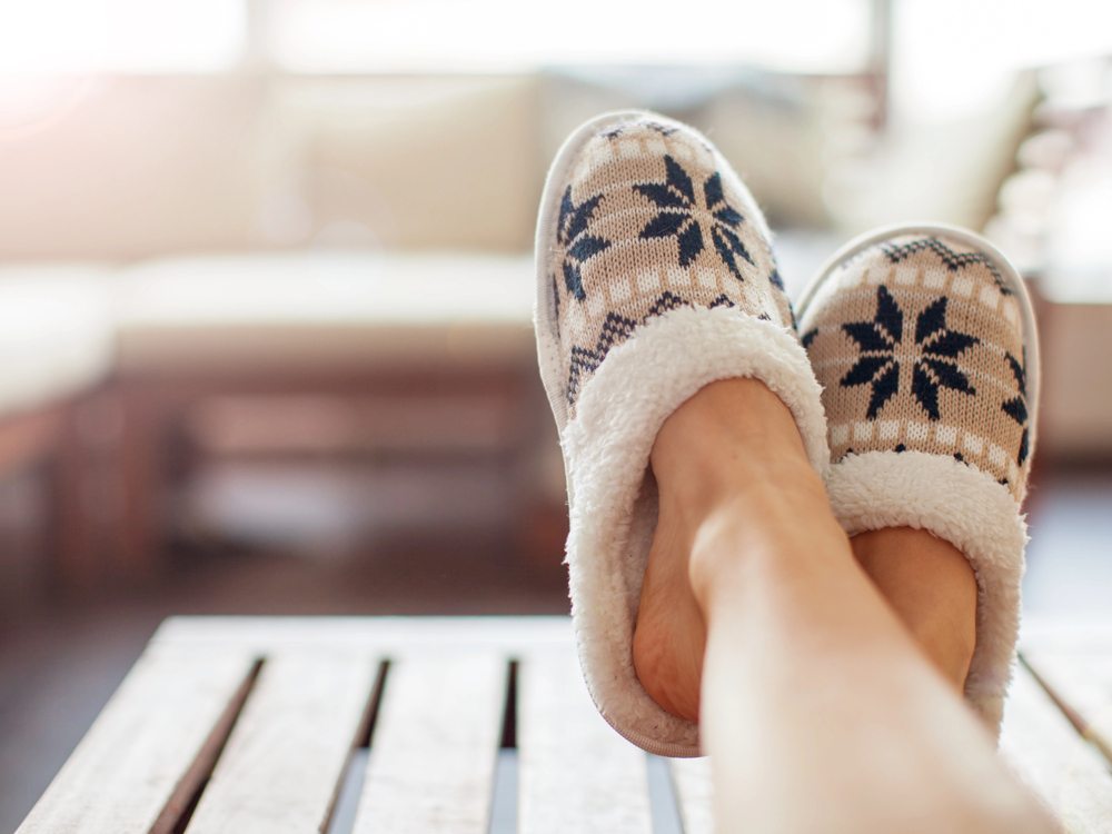 A person relaxes with their feet up on a wooden table, wearing cosy slippers with a beige, white, and navy blue snowflake pattern. The background shows a bright, softly focussed living room.