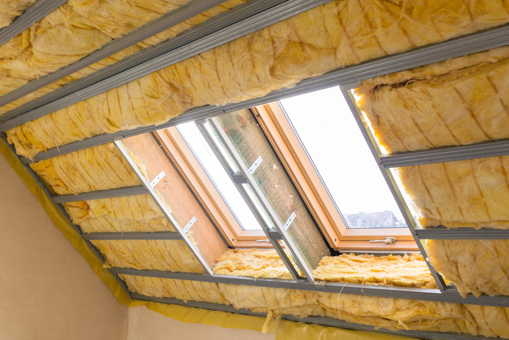 Attic ceiling under construction, showing exposed yellow fibreglass insulation and metal framing, with a wooden-framed rooflight window letting in natural light.