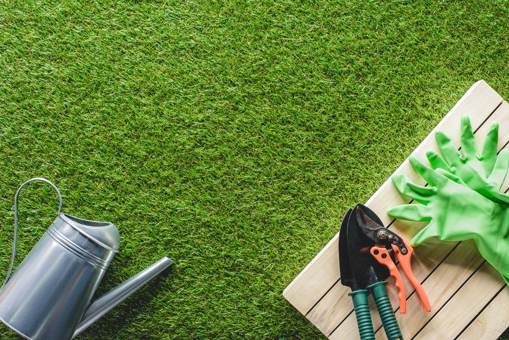 A metal watering can, green gardening gloves, and secateurs are arranged on a wooden board on a grassy surface, with ample open space on the left side.