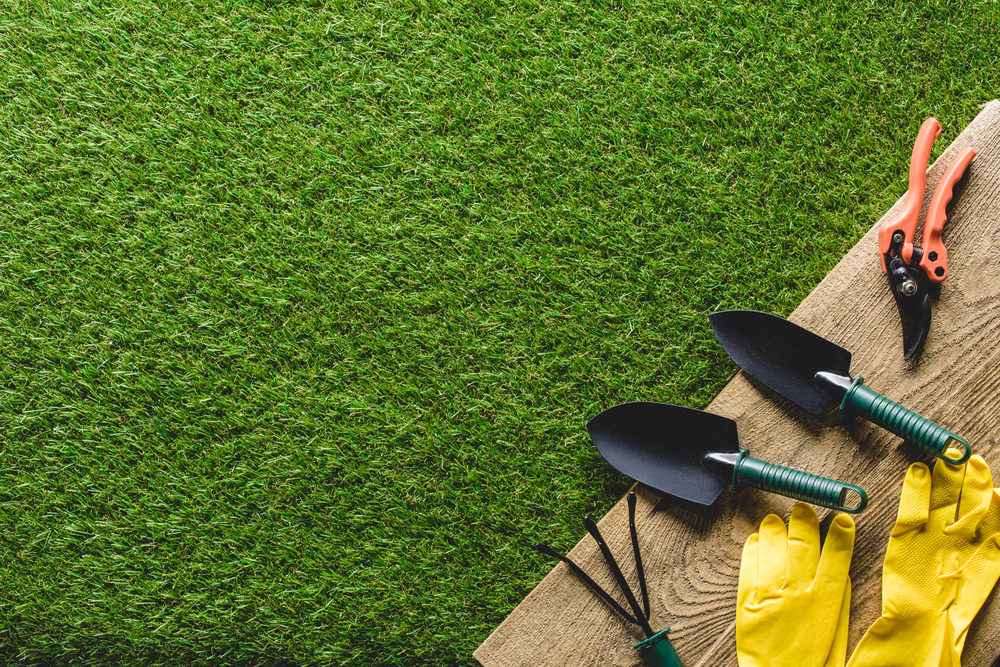 Gardening tools including two trowels, a hand rake, secateurs, and yellow gloves are arranged on a wooden plank next to a patch of green grass, with ample space on the left side.