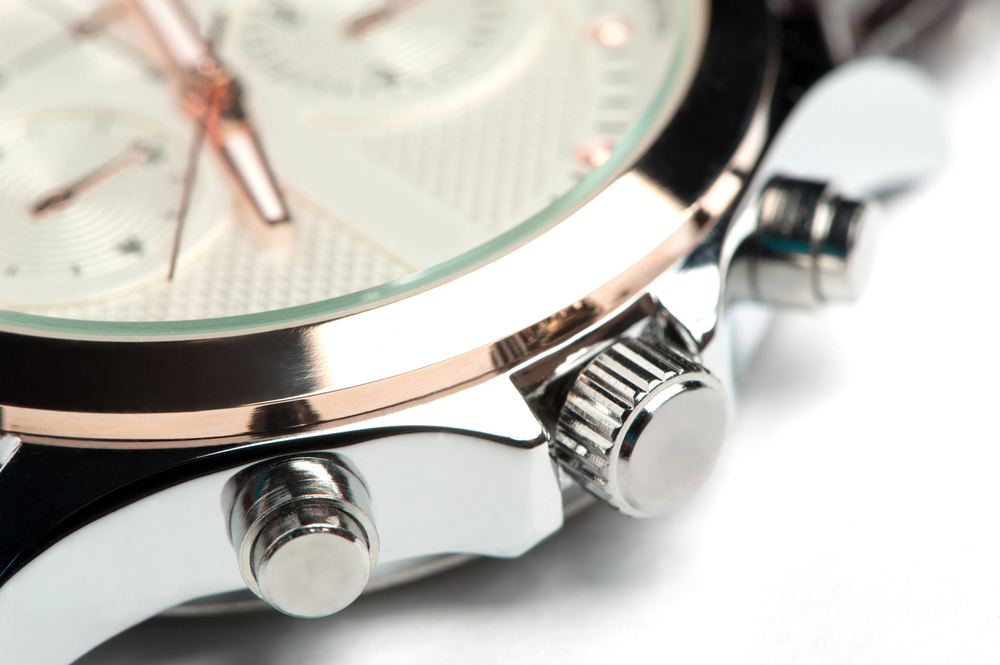 Close-up of a silver wristwatch showing the crown and two push-buttons on the side, with part of the round, textured watch face and glass visible.