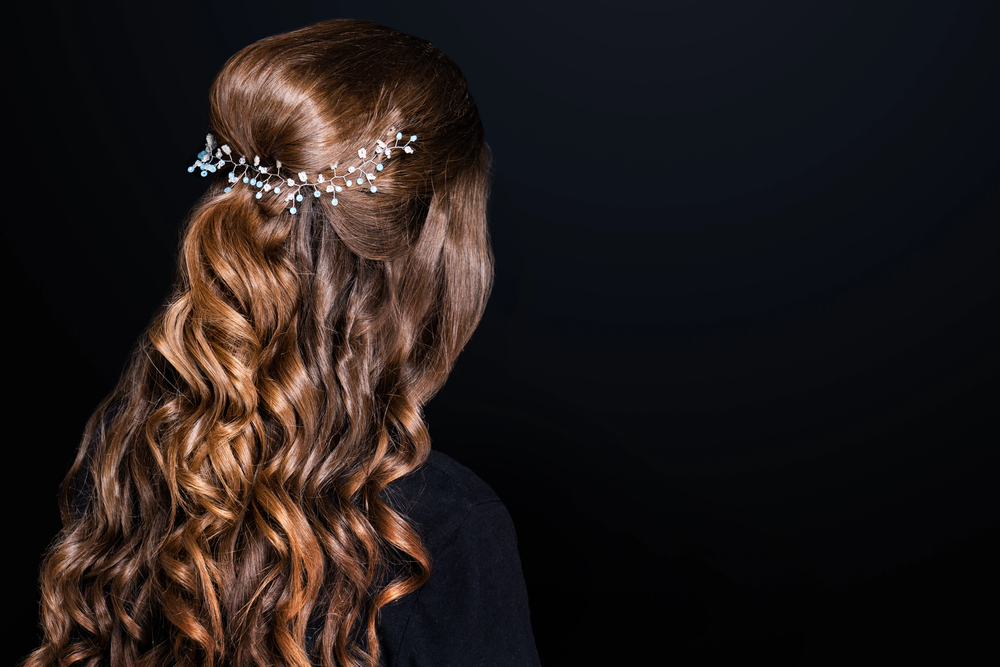 A woman with long, wavy brown hair styled half up, accessorised with a decorative hairpiece featuring small pearls and crystal-like beads, is photographed from behind against a black background.