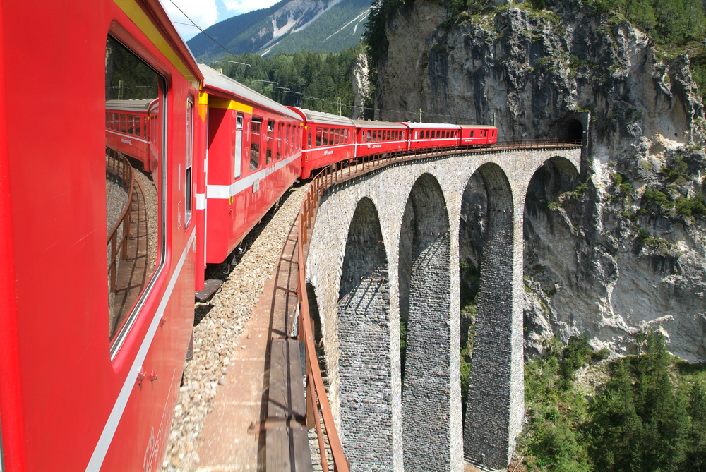 A bright red passenger train travels across a tall stone viaduct, curving along a mountainside surrounded by rocky cliffs and green forest.