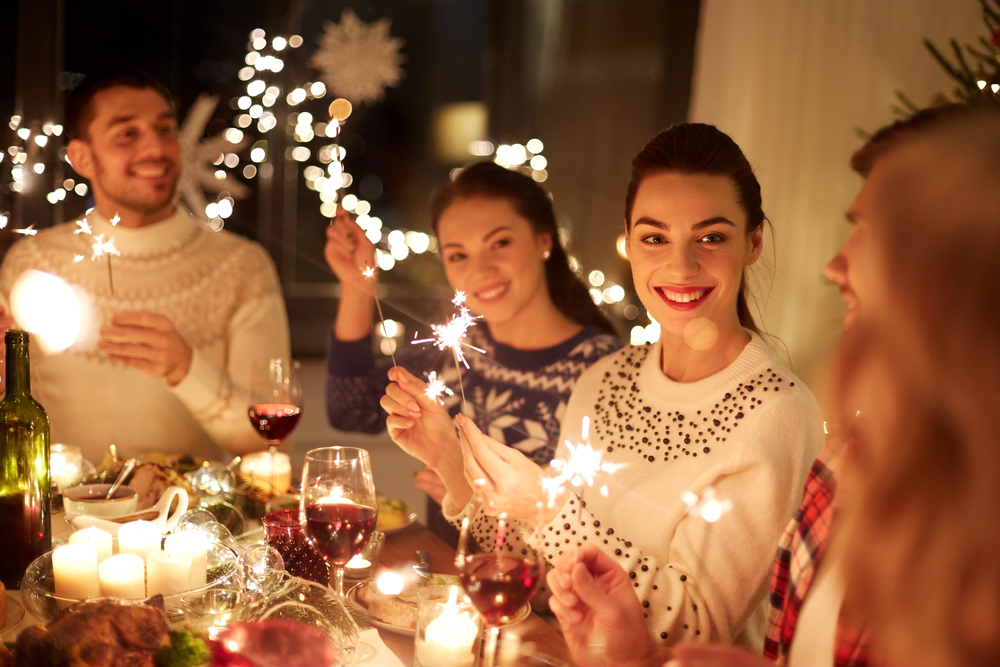 Four people sit around a festive dinner table, holding sparklers and smiling. The table is set with food, candles, and glasses of red wine, with fairy lights glowing in the background.