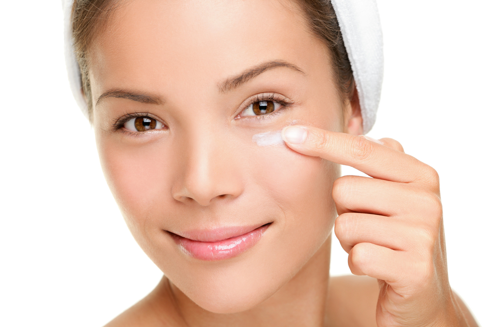 A woman with a towel wrapped around her head is smiling and applying a small amount of cream under her eye with her finger against a white background.