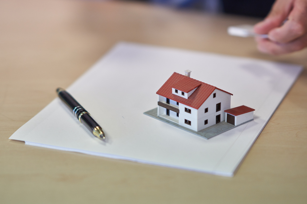 A model house with a red roof sits on a stack of papers next to a black pen on a desk, with a person's hand visible in the background.