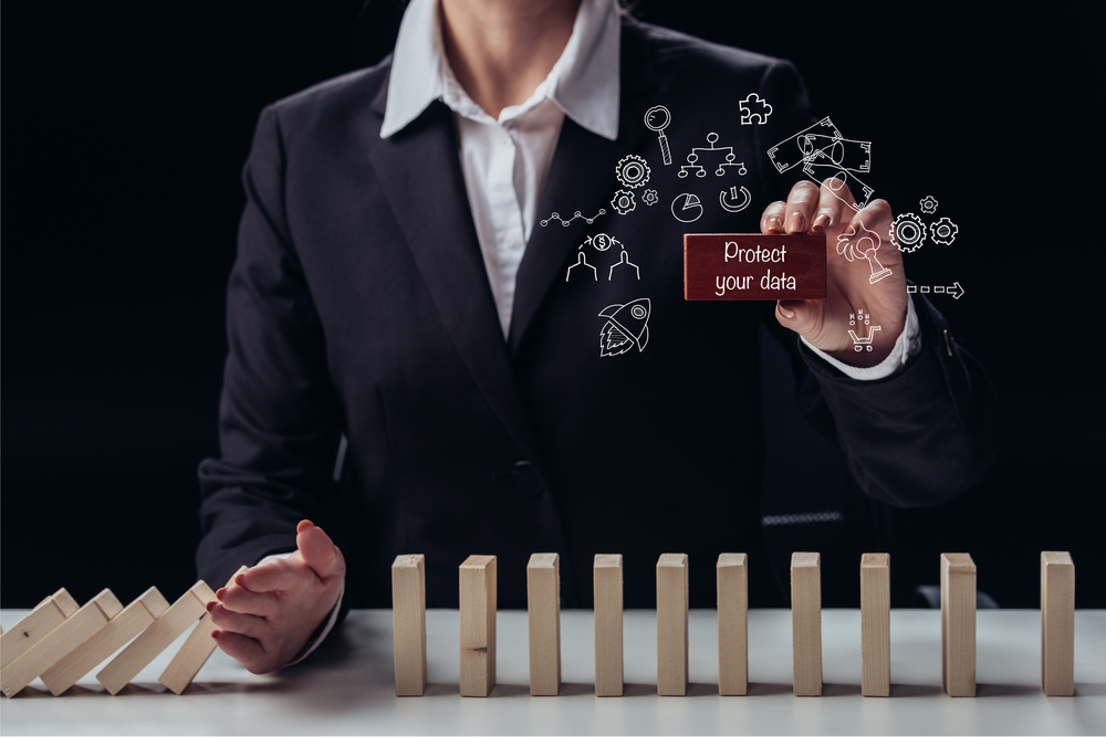 A person in a suit stops falling wooden dominoes with one hand while holding a card that says Protect your data with tech icons above it, symbolising data protection and cybersecurity.