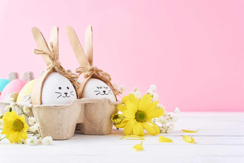 Two Easter eggs decorated as bunnies with drawn faces, wooden ears, and twine bows sit in a carton. Yellow daisies and small white flowers are arranged beside them against a pink background.