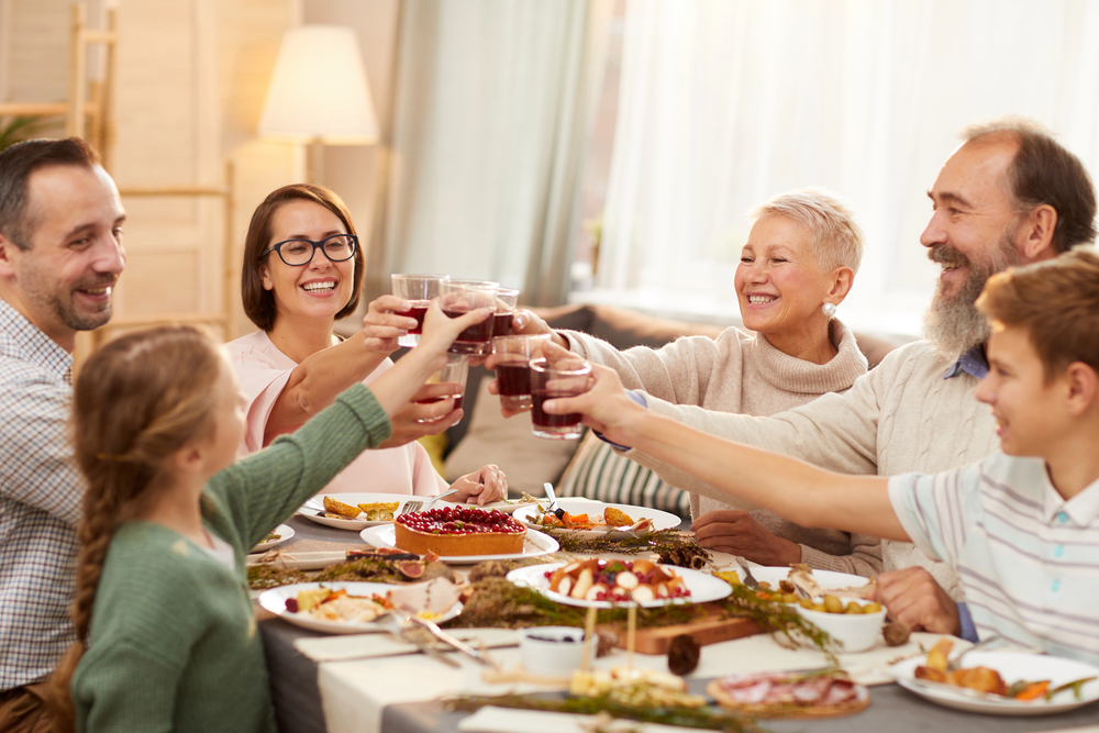 A multigenerational family sits around a dining table, smiling and raising glasses in a toast over a festive meal with assorted dishes and decorations.