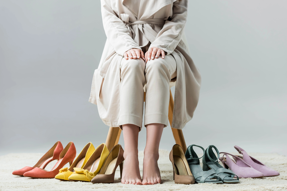 A person in a beige outfit sits barefoot on a stool, with various pairs of colourful high-heeled shoes arranged on the floor in front of them.