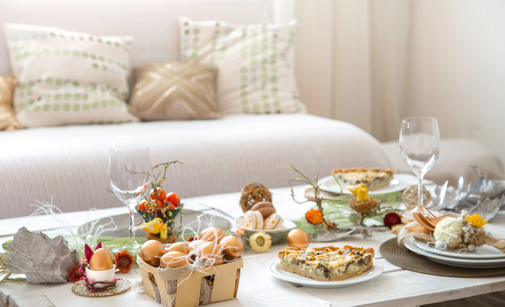 A bright, elegant table is set with crockery, eggs in a basket, quiche, wine glasses, and spring-themed decorations, in front of a white sofa with patterned cushions.