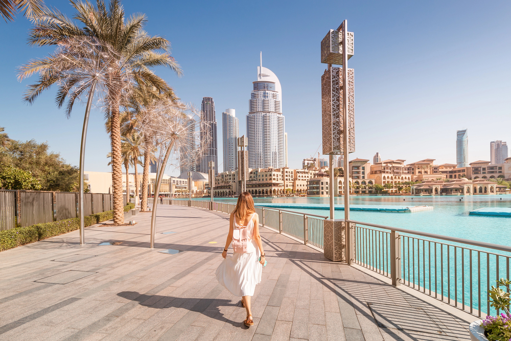 A woman in a white dress walks along a sunny waterfront promenade with palm trees, modern skyscrapers, and turquoise water in a cityscape, likely Dubai.