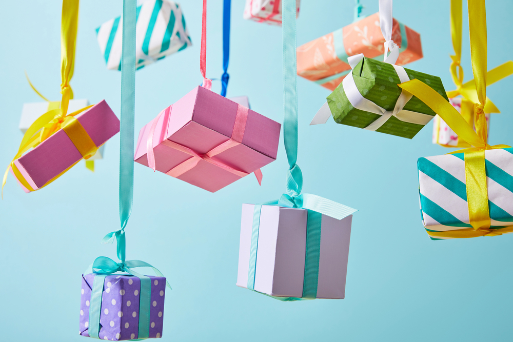 Colourful gift boxes with ribbons hang from above against a light blue background. The boxes are wrapped in various patterns, including stripes, polka dots, and solids, creating a festive and cheerful scene.