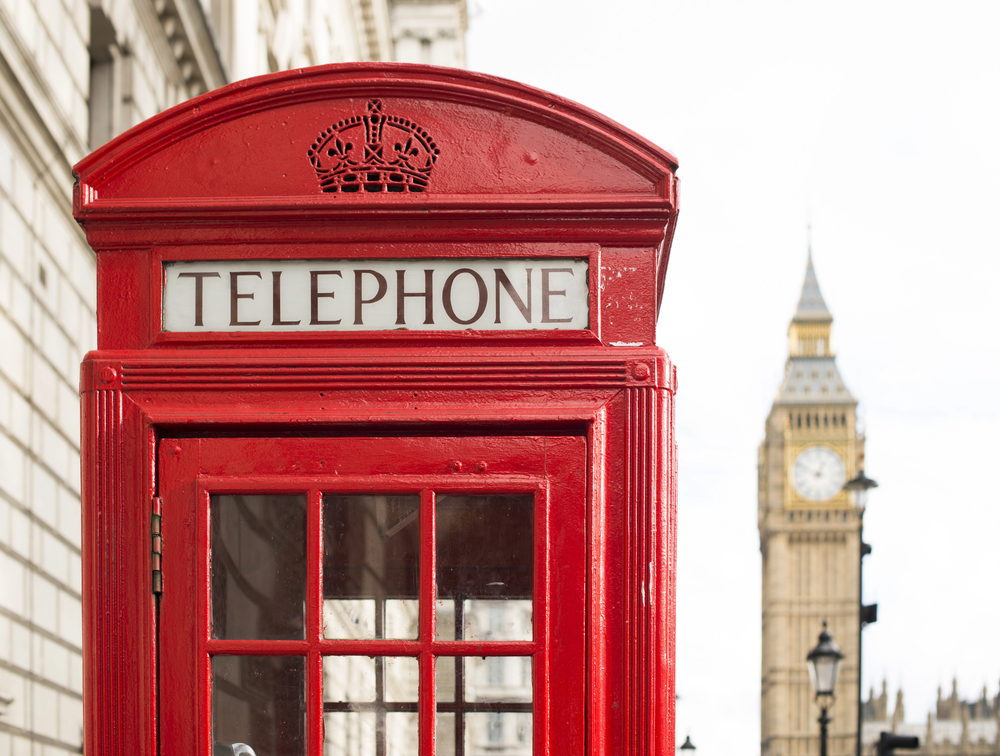 A classic red British telephone box stands in the foreground, with the iconic clock tower of Big Ben blurred in the background on a bright, clear day in London.
