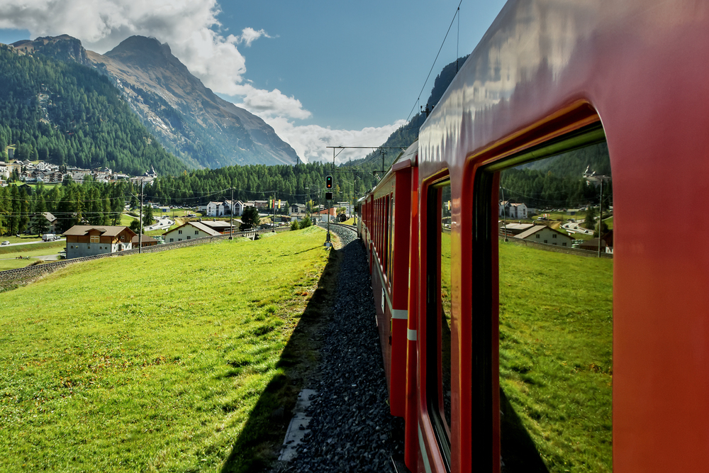 A red train travels through a lush green valley with mountain peaks in the background, passing small houses and pine forests under a partly cloudy sky.