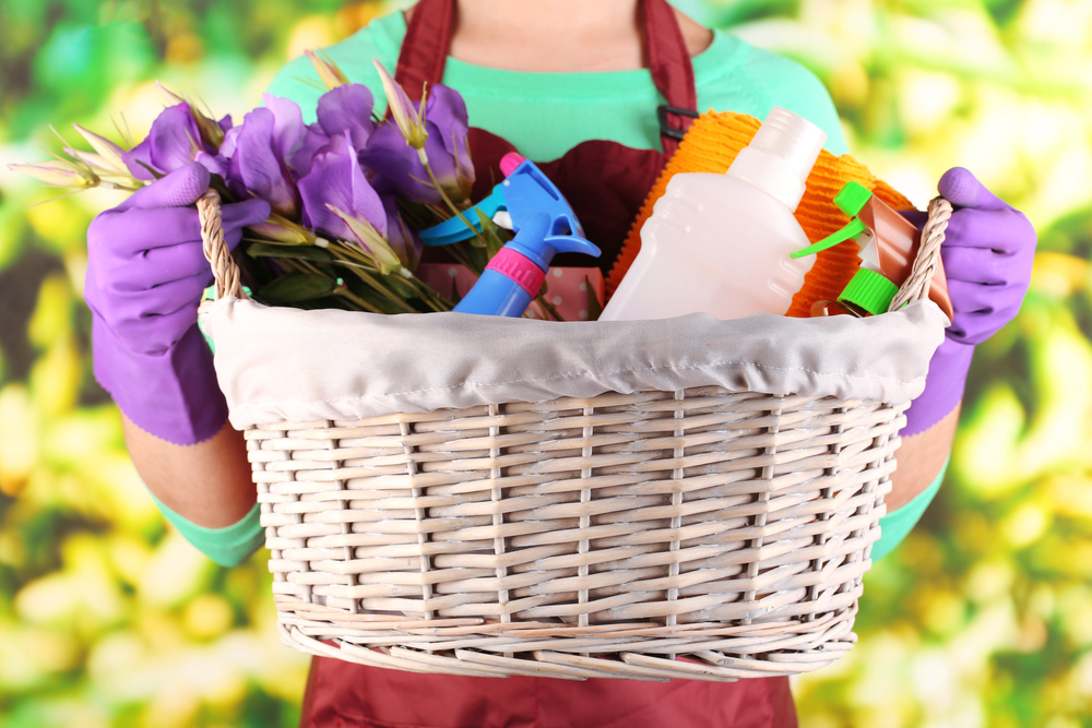 A person wearing purple gloves holds a white wicker basket filled with purple flowers, cleaning spray bottles, and yellow cleaning cloths. The background is green and yellow with a blurred, leafy pattern.