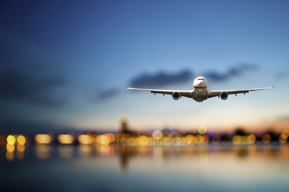 A commercial aeroplane flies low over water towards the camera at dusk, with a colourful sky and city lights reflecting on the water in the background.