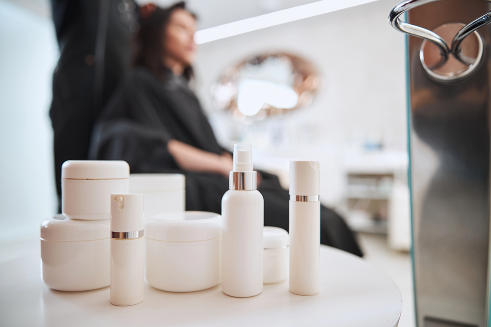 Several white cosmetic containers are arranged on a table in the foreground, while a person with dark hair sits blurred in the background, suggesting a salon or beauty treatment setting.