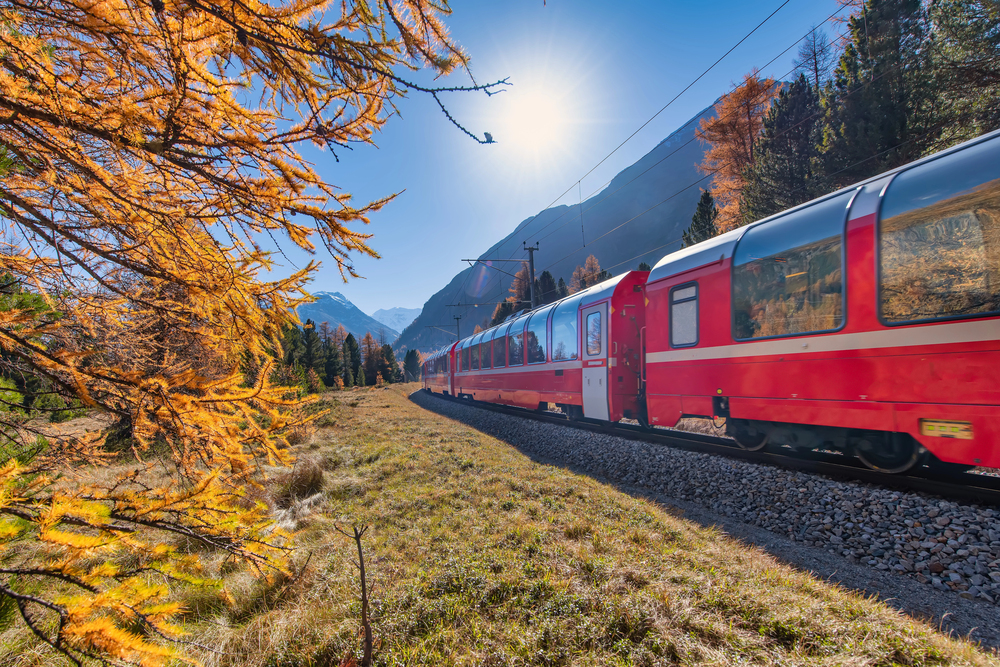 A red passenger train travels through a scenic mountain landscape with autumn trees, under a clear blue sky and bright sun, with snow-capped peaks visible in the background.