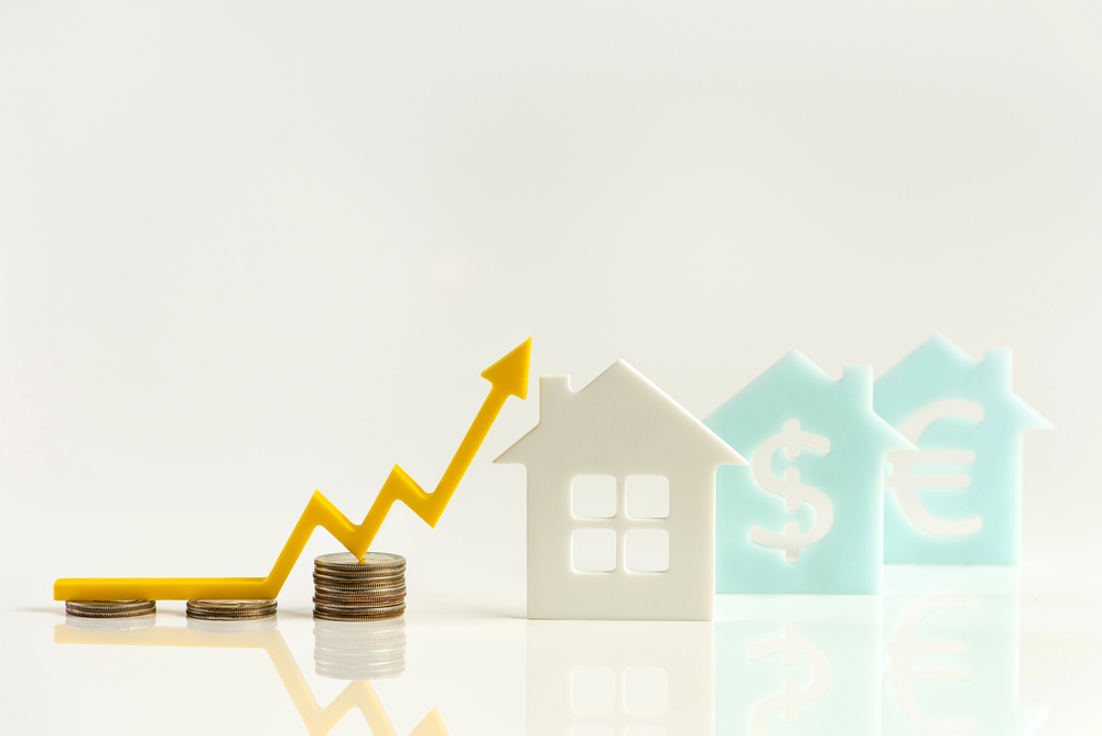 A yellow upward arrow rests on stacks of coins, next to white and blue house-shaped figures with dollar and euro symbols, representing rising property or housing market values.