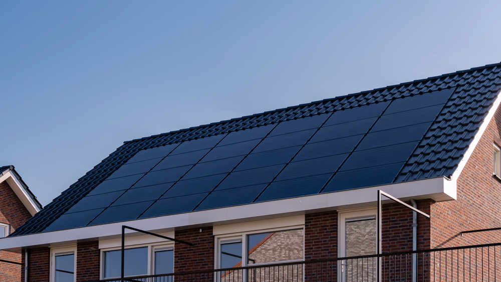 A house with a sloping roof covered in black solar panels and dark roof tiles, set against a clear blue sky.