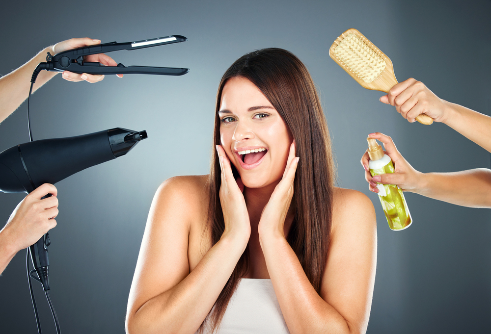 A smiling woman with straight brown hair is surrounded by hands holding a hairdryer, hair straightener, hairbrush, and a spray bottle, suggesting a hair care or beauty routine against a grey background.