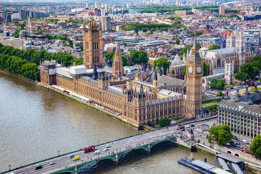 Aerial view of the Palace of Westminster and Big Ben by the River Thames in London, with Westminster Bridge and city buildings in the background.
