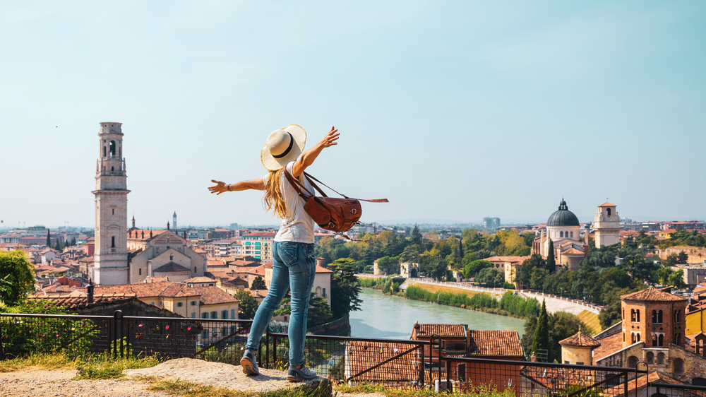 A woman with long blonde hair and a hat stands on a hill overlooking a scenic city with historic buildings, a river, and church towers, spreading her arms wide in joy under a clear blue sky.
