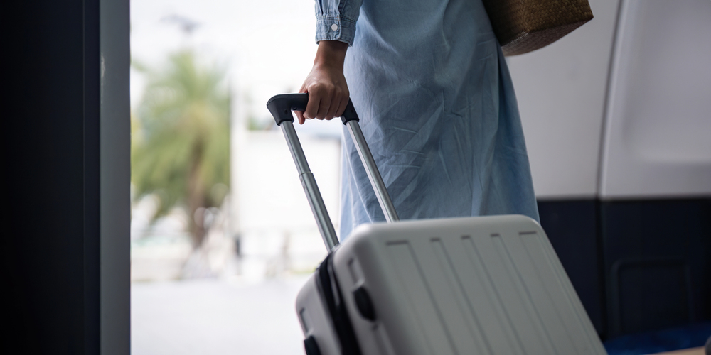 A person wearing a light blue dress holds the handle of a white rolling suitcase, standing near a doorway with an outdoor scene visible in the background.