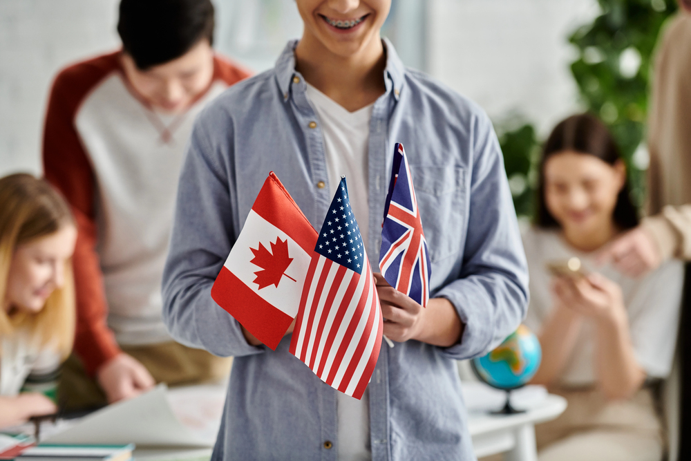 A person holds small Canadian, American, and British flags. Other people are in the background, some working at a table and one using a mobile phone. The setting appears to be a classroom or study space.