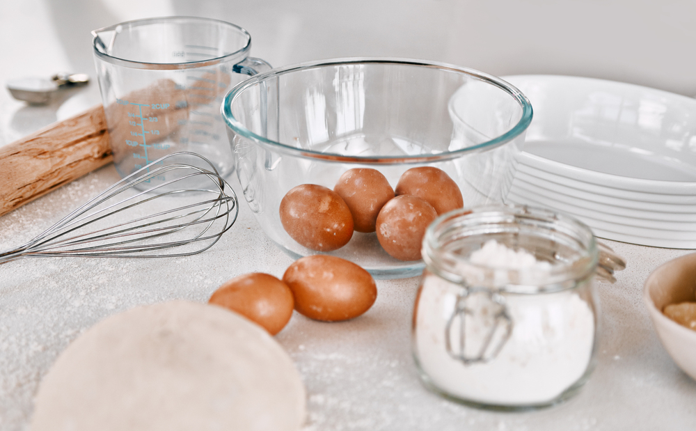 A kitchen worktop with baking ingredients and tools: brown eggs in a glass bowl, a whisk, a rolling pin, a jar of flour, a measuring jug, a stack of plates, and a circle of dough on a floured surface.