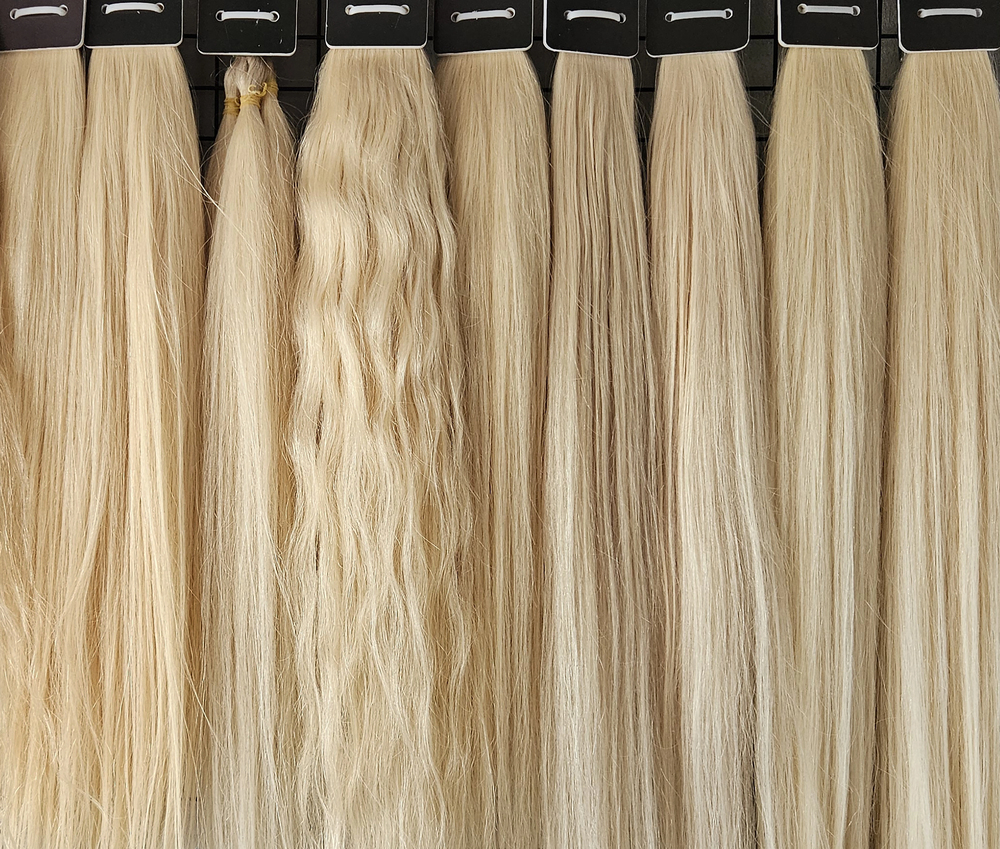Several samples of blonde hair extensions, each with slightly different textures and shades, are displayed vertically in a row, attached to black holders at the top.