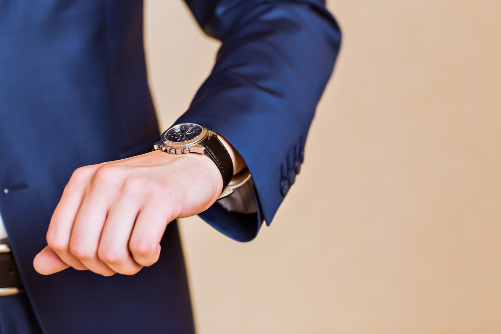 A person in a blue suit is checking the time on a wristwatch, with only their hand, wrist, and part of their torso visible against a plain background.