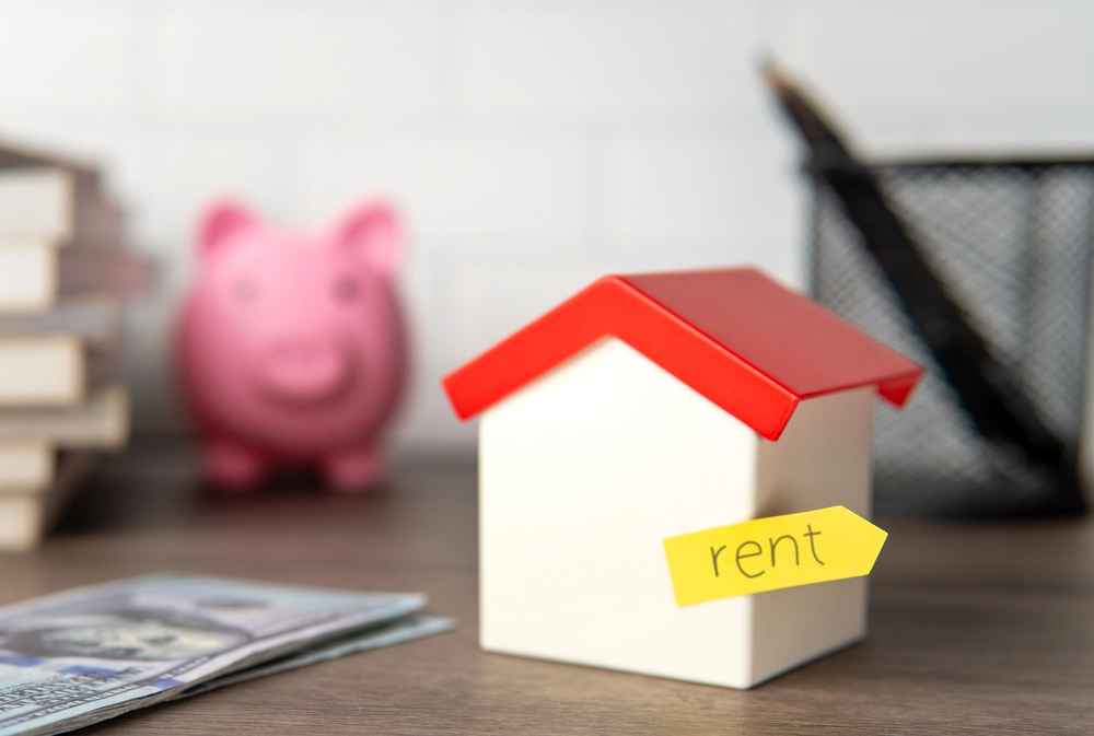 A small model house with a red roof and a yellow tag labelled rent sits on a desk. In the background, there is a pink piggy bank, a stack of books, and a pen holder. Money is in the foreground.