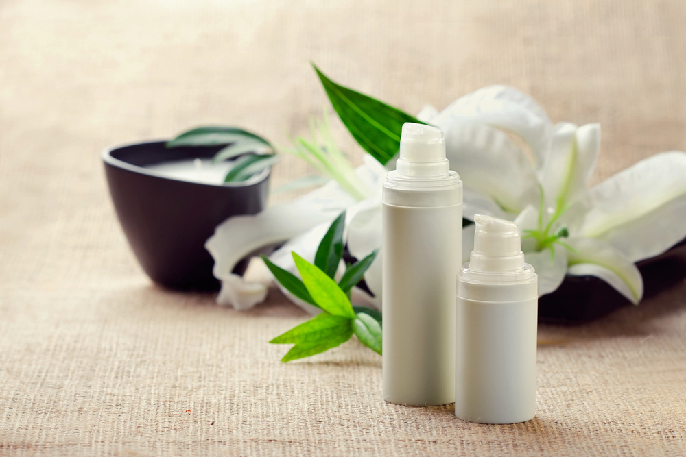 Two white cosmetic pump bottles are placed on a beige fabric surface, with green leaves, a white lily flower, and a black bowl candle in the background, creating a spa-like and calming atmosphere.