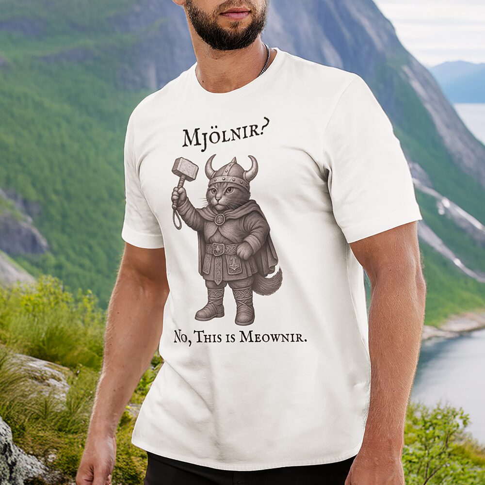 A man stands outdoors wearing a white T-shirt featuring an illustration of a Viking cat holding a hammer with the caption: Mjölnir? No, This is Meownir. Mountains and greenery are in the background.