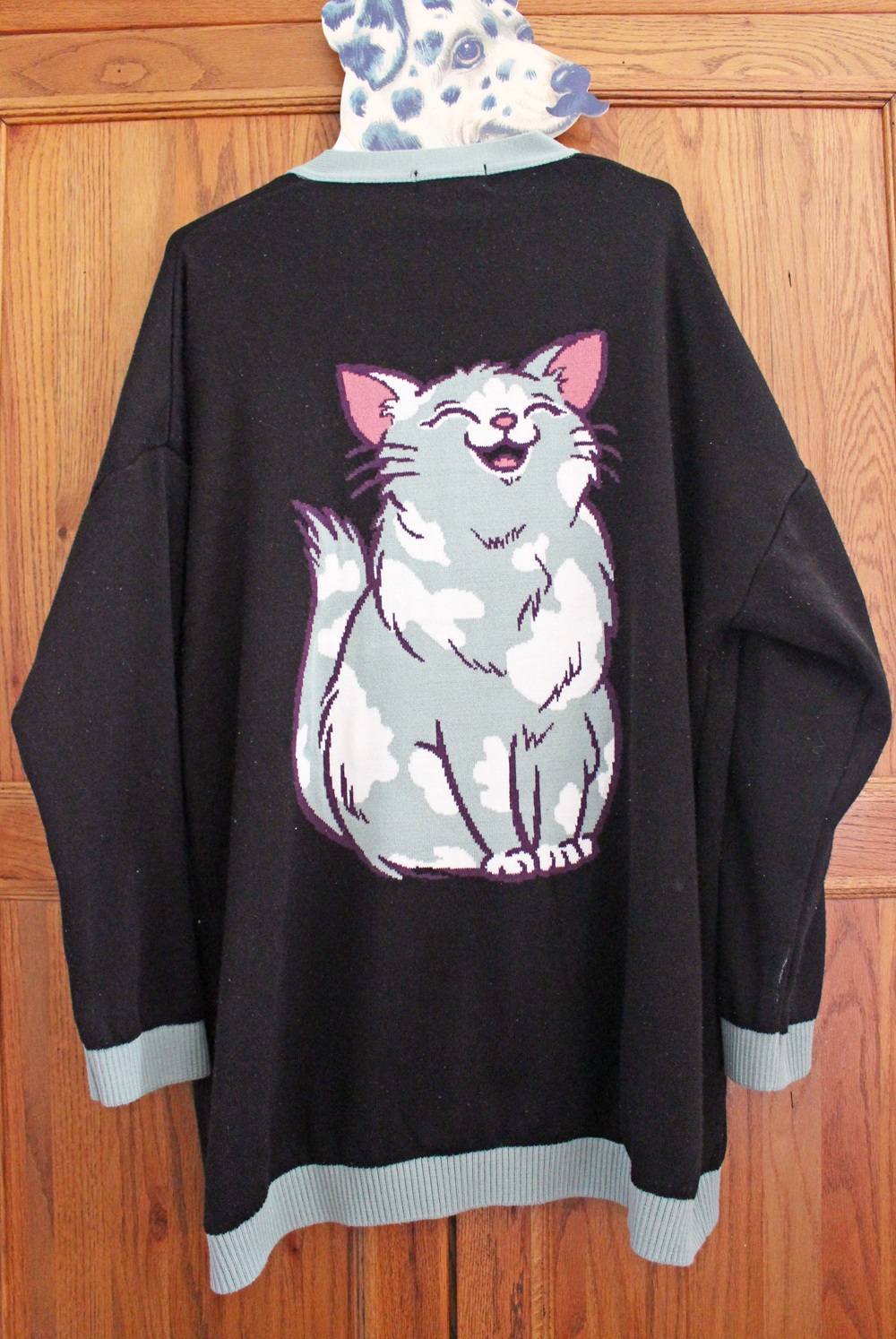 A black jumper with light blue trim hangs on a wooden door. The back of the jumper features a large, cartoon-style image of a smiling white cat with light blue and lavender shading.