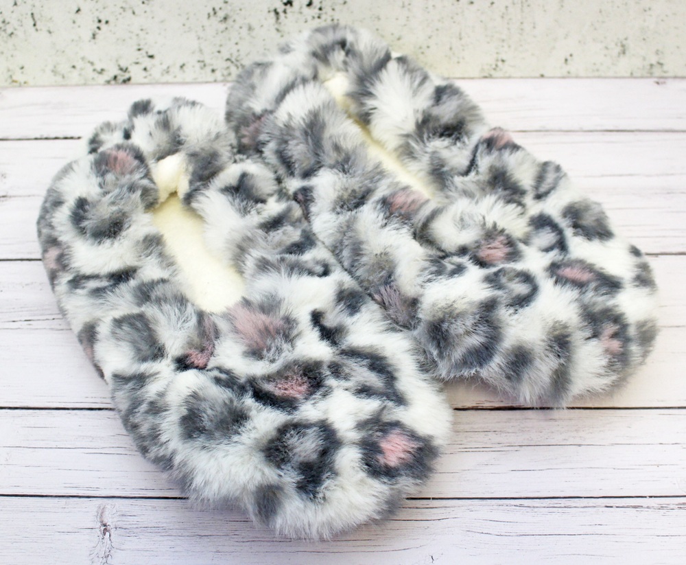 A pair of fuzzy, faux fur slippers with a white base and grey and pink leopard-like spots—so comfy they're sure to inspire repeat purchases—placed on a light wooden surface against a speckled white background.